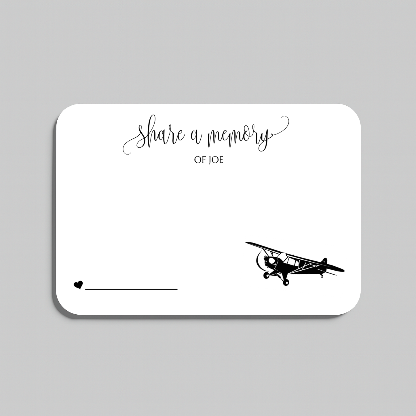 A white rectangular memorial card with rounded corners, featuring the script text 'share a memory' at the top, a small heart and line at the bottom left, and a black vintage airplane illustration at the bottom right. The card is shown on a light gray background.