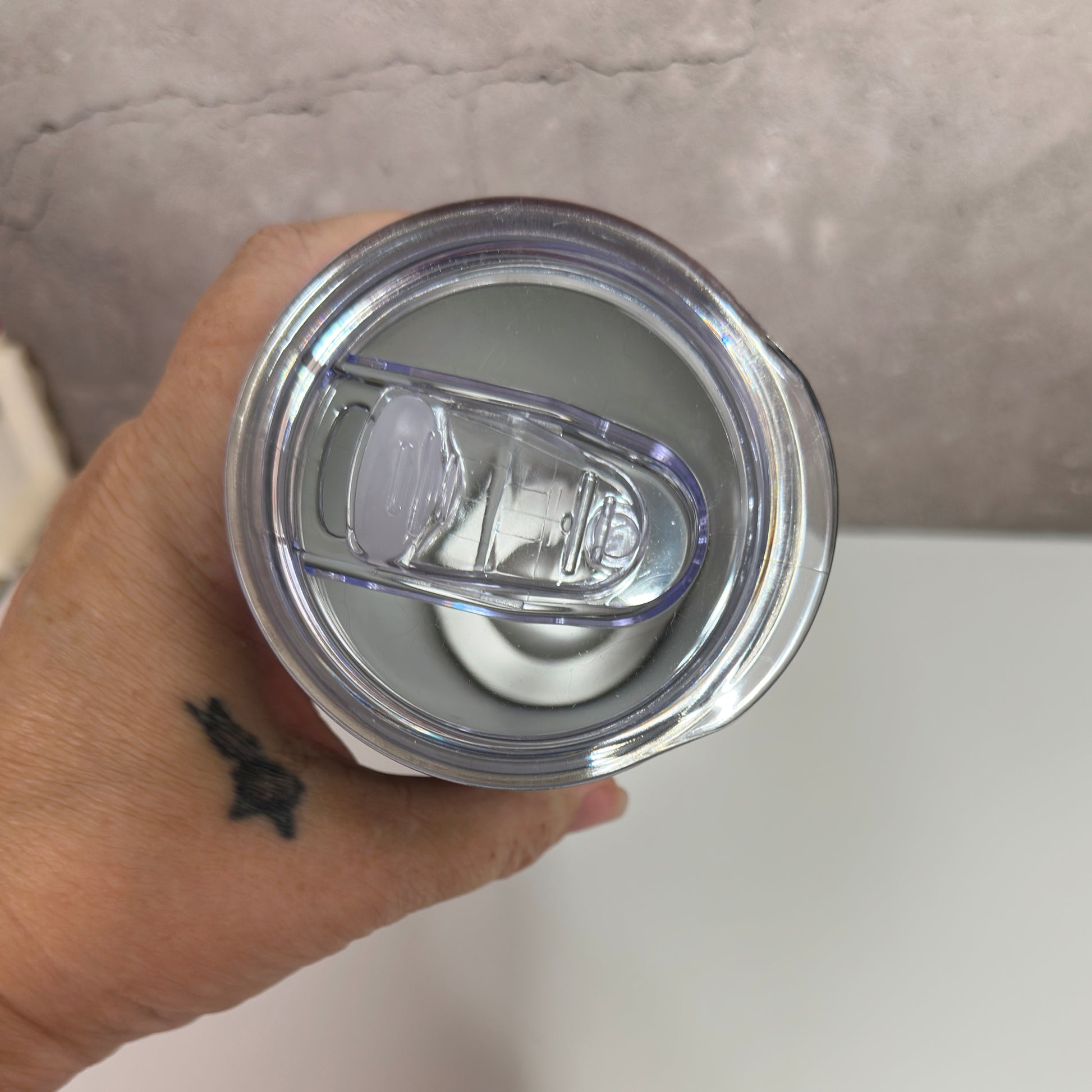 Hand holding a clear tumbler with a silver lid against a neutral background