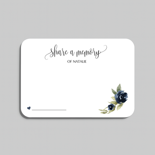A rectangular white memorial card with rounded corners, featuring the script text 'share a memory  at the top, a small line for writing at the bottom left, and a watercolor floral design in navy blue and green at the bottom right. The card is centered on a light gray background.