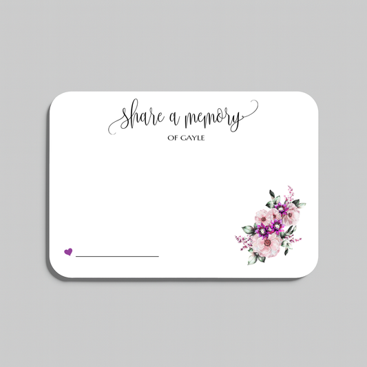 A rectangular white memorial card with rounded corners, featuring the script text 'share a memory' at the top, a small purple heart and line in the lower left, and a watercolor floral bouquet in pink, purple, and green in the lower right. The card is shown on a plain light gray background.