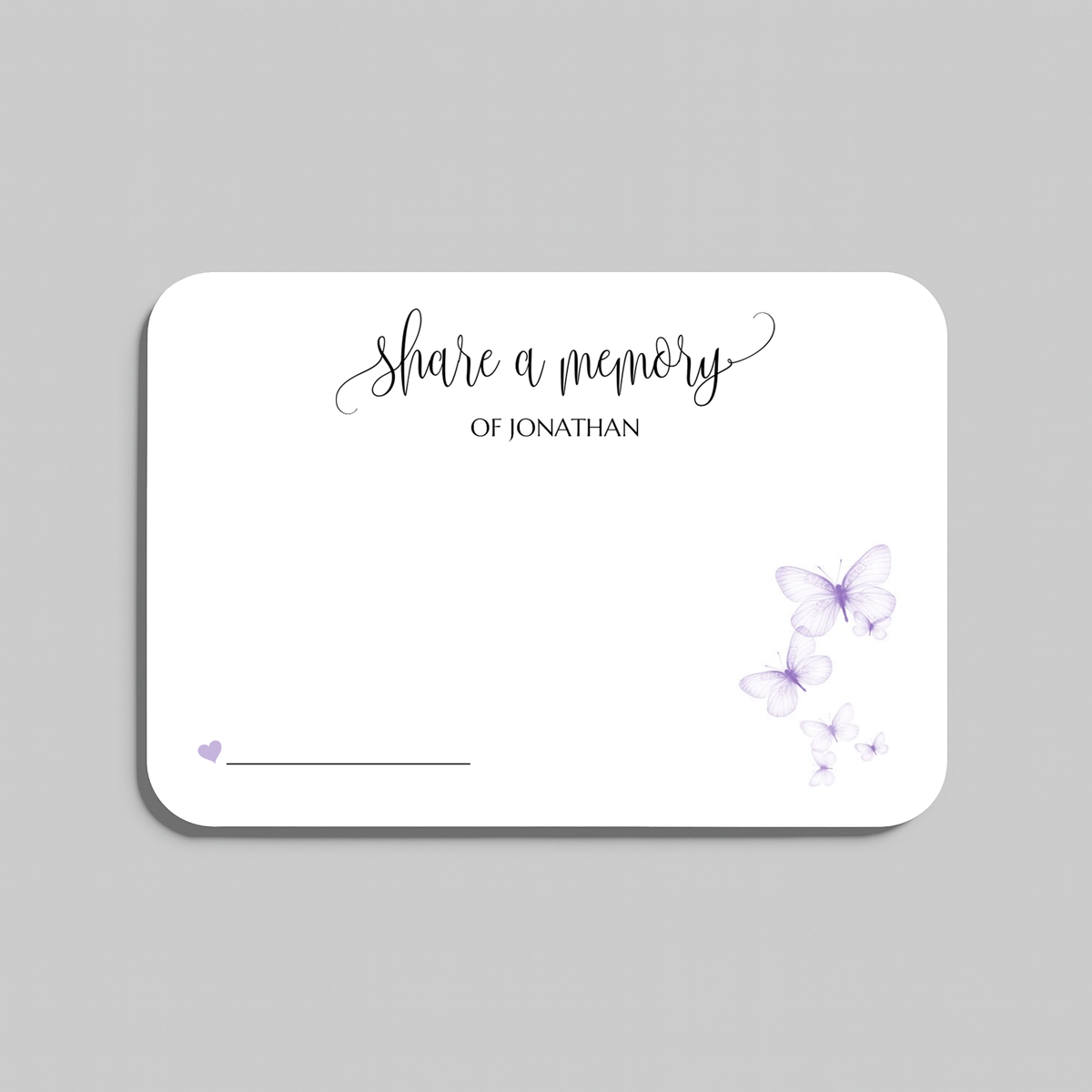 A white rectangular memorial card with rounded corners, featuring the printed text 'Share a memory' in black script and serif fonts, decorated with light purple watercolor butterflies and a small heart near the bottom left, shown on a neutral gray background.