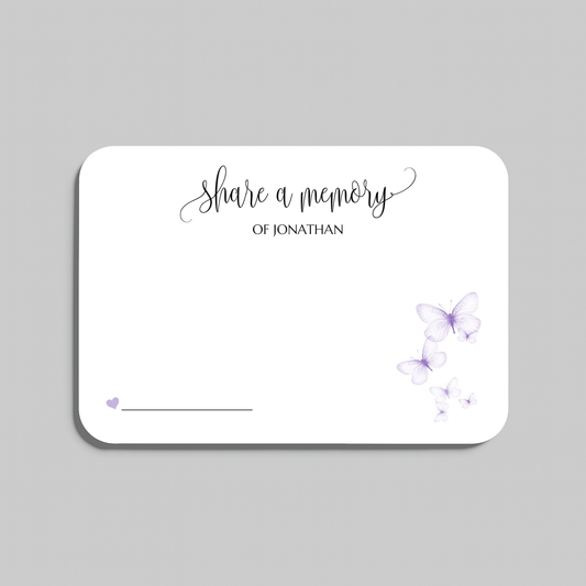 A white rectangular memorial card with rounded corners, featuring the printed text 'Share a memory' in black script and serif fonts, decorated with light purple watercolor butterflies and a small heart near the bottom left, shown on a neutral gray background.