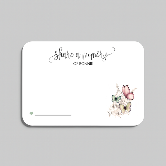 A white rectangular memorial card with rounded corners, featuring the script text 'share a memory' and a small floral-and-butterfly illustration in the lower right. The card is shown on a light gray background.