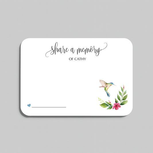 A white memorial guest book card with rounded corners, featuring the script text 'Share a memory' and a watercolor illustration of a hummingbird with green leaves and pink flowers in the lower right corner, shown on a neutral gray background.