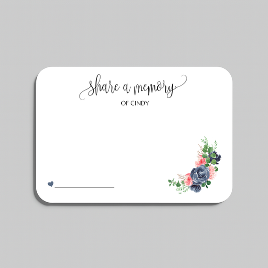 A white rectangular memorial card with rounded corners, featuring the script text 'share a memory' at the top, a blank line at the bottom left, and a small floral arrangement in the lower right corner. The card is shown on a neutral gray background.