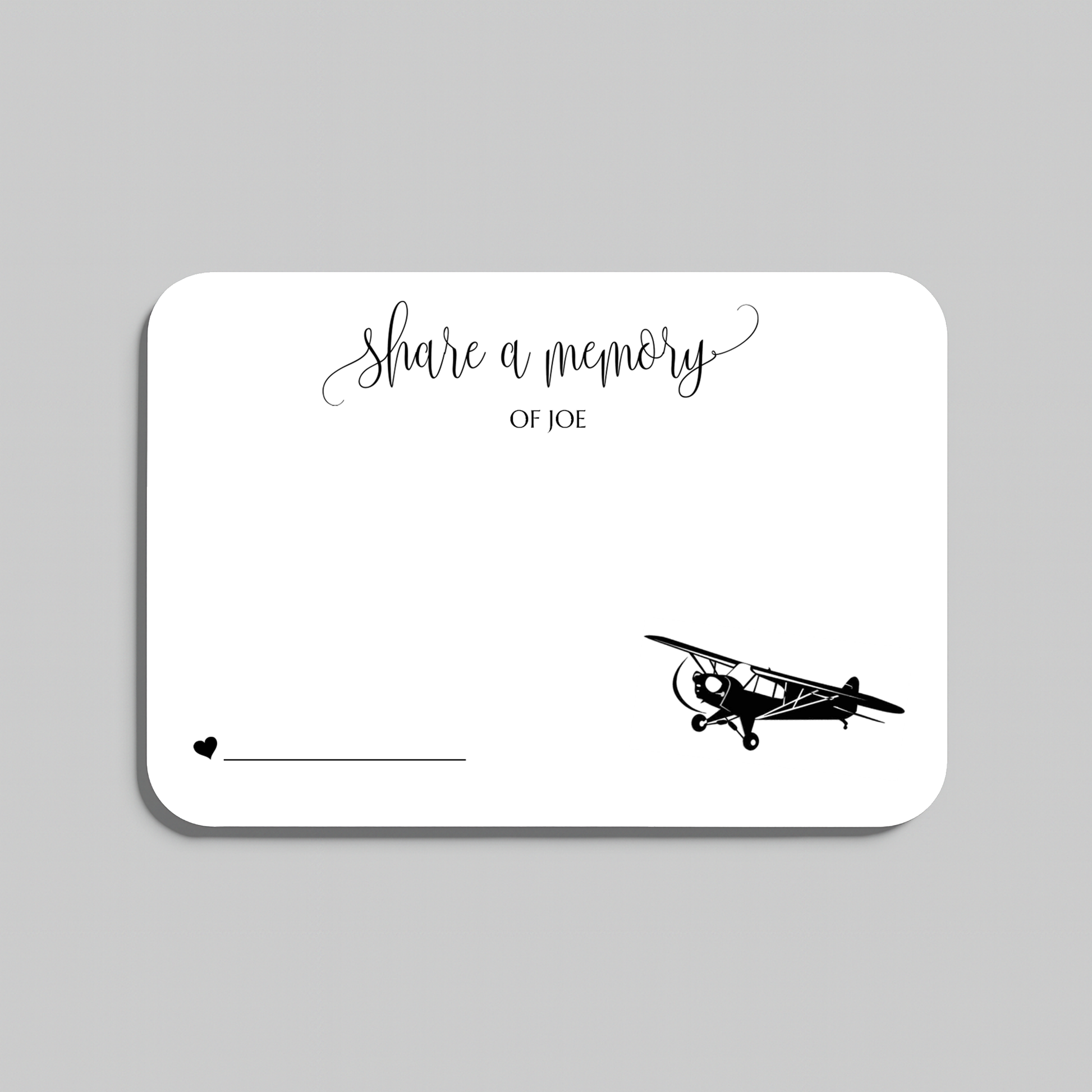 A white rectangular memorial card with rounded corners, featuring the script text 'share a memory' at the top, a small heart and line at the bottom left, and a black vintage airplane illustration at the bottom right. The card is shown on a light gray background.