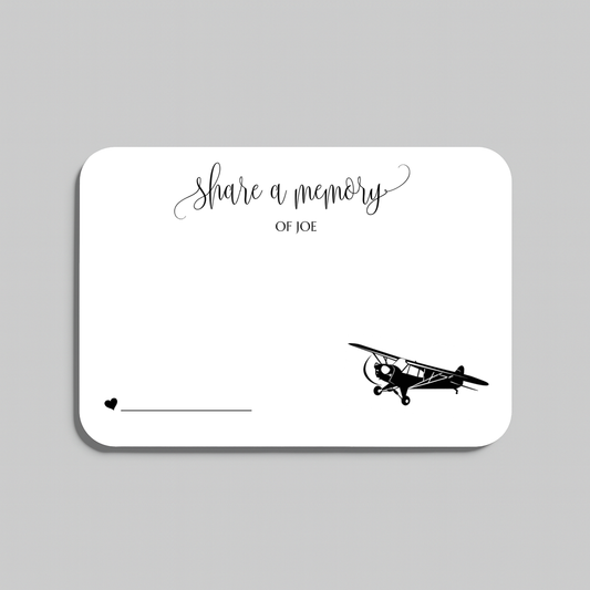 A white rectangular memorial card with rounded corners, featuring the script text 'share a memory' at the top, a small heart and line at the bottom left, and a black vintage airplane illustration at the bottom right. The card is shown on a light gray background.