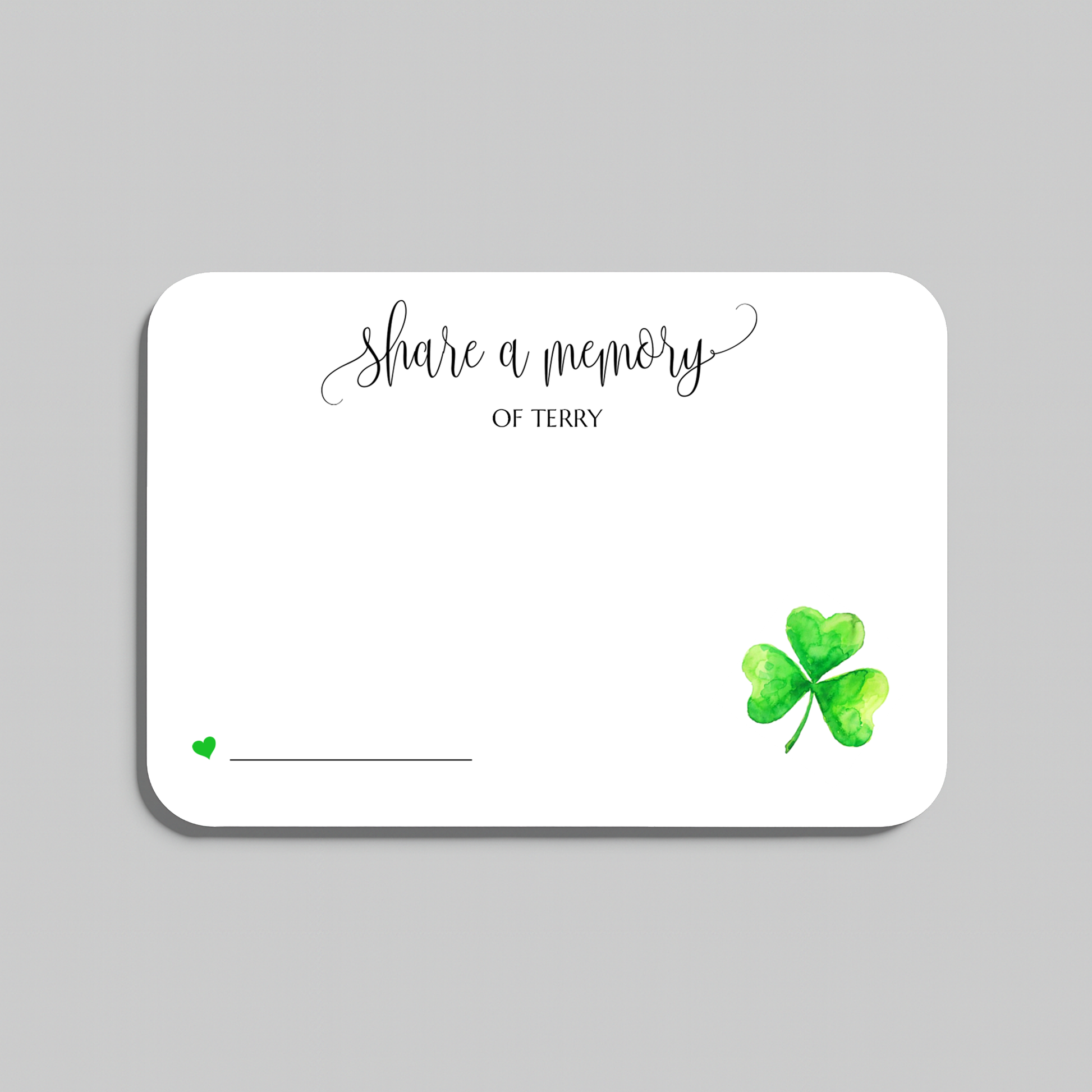 A white rectangular memorial card with rounded corners, featuring the script text 'share a memory' at the top, a small green heart and line near the bottom left, and a green watercolor shamrock in the bottom right. The card is shown on a light gray background.