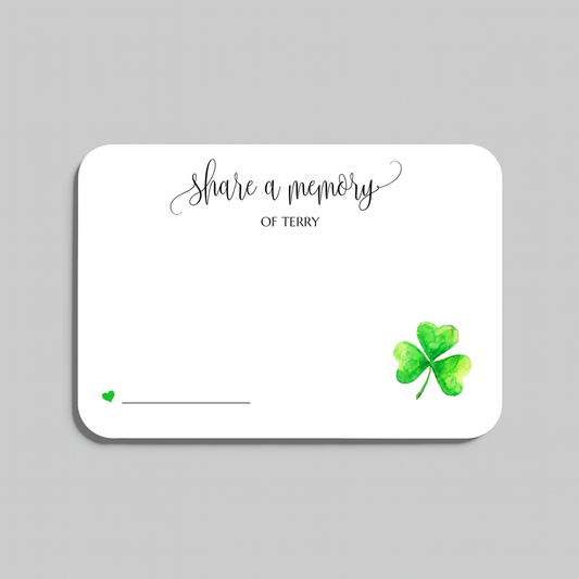 A white rectangular memorial card with rounded corners, featuring the script text 'share a memory' at the top, a small green heart and line near the bottom left, and a green watercolor shamrock in the bottom right. The card is shown on a light gray background.