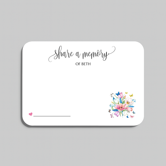A white rectangular memorial card with rounded corners, featuring the script text 'share a memory' at the top, a small pink heart and line in the lower left, and a colorful floral bouquet illustration in the lower right. The card is shown on a light gray background.