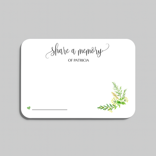 A white rectangular memorial card with rounded corners, featuring the script text 'share a memory' at the top, a small green heart and line at the bottom left, and a delicate green and yellow floral illustration in the lower right corner. The card is centered on a solid gray background.