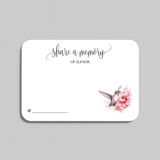 A rectangular white memorial card with rounded corners, featuring the script text 'share a memory' at the top, a small heart and line in the lower left, and a watercolor hummingbird with pink flowers in the lower right. The card is shown on a light gray background.